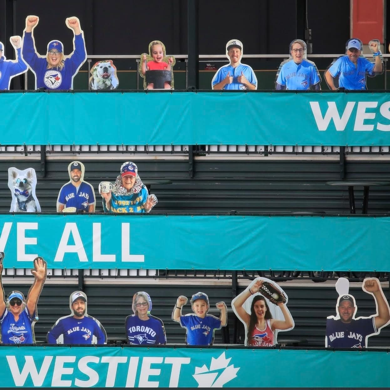 Cardboard cutouts of Toronto Blue Jays fans, including adults, children, and pets, are arranged in stadium seats. The cutouts wear Blue Jays apparel and appear to be cheering. Large teal banners with white text, such as WESTJET and WE ALL, are visible in the background, creating a festive and supportive atmosphere that represents fan presence during a game.